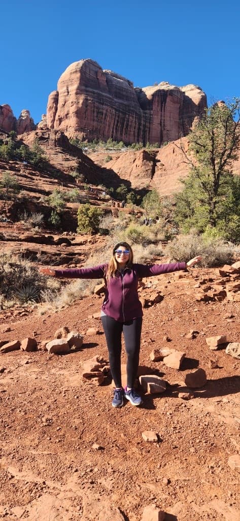 Woman in a purple jacket standing with arms open before Sedona red rock cliffs
